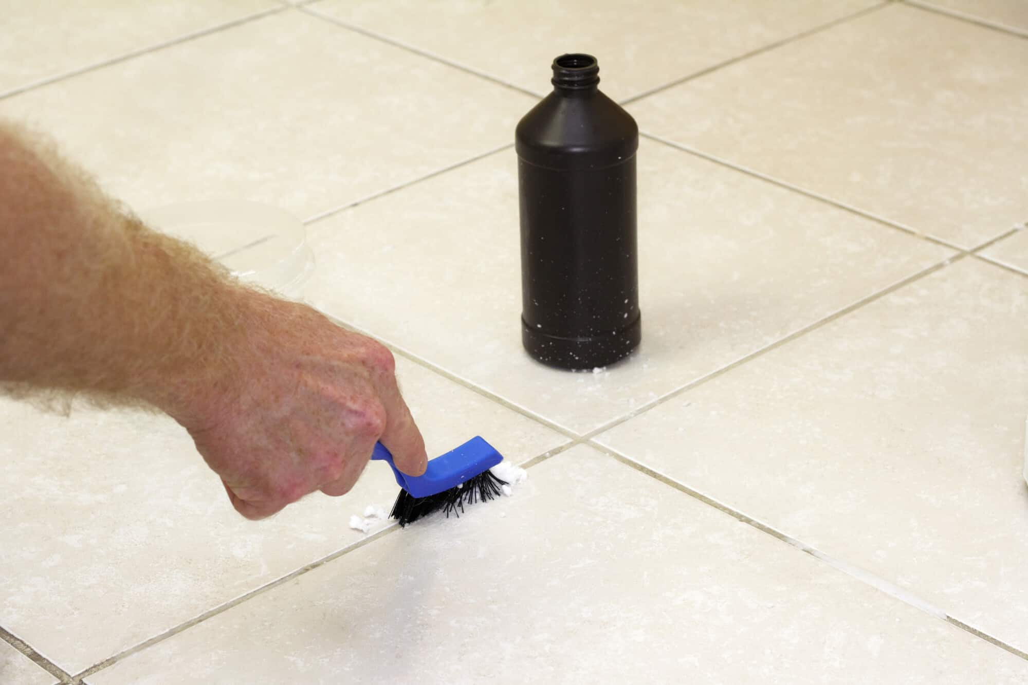 Hand with a blue handled black scrub brush cleaning grout with baking soda and peroxide. Floor grout tile being cleaned with baking soda and hydrogen peroxide.