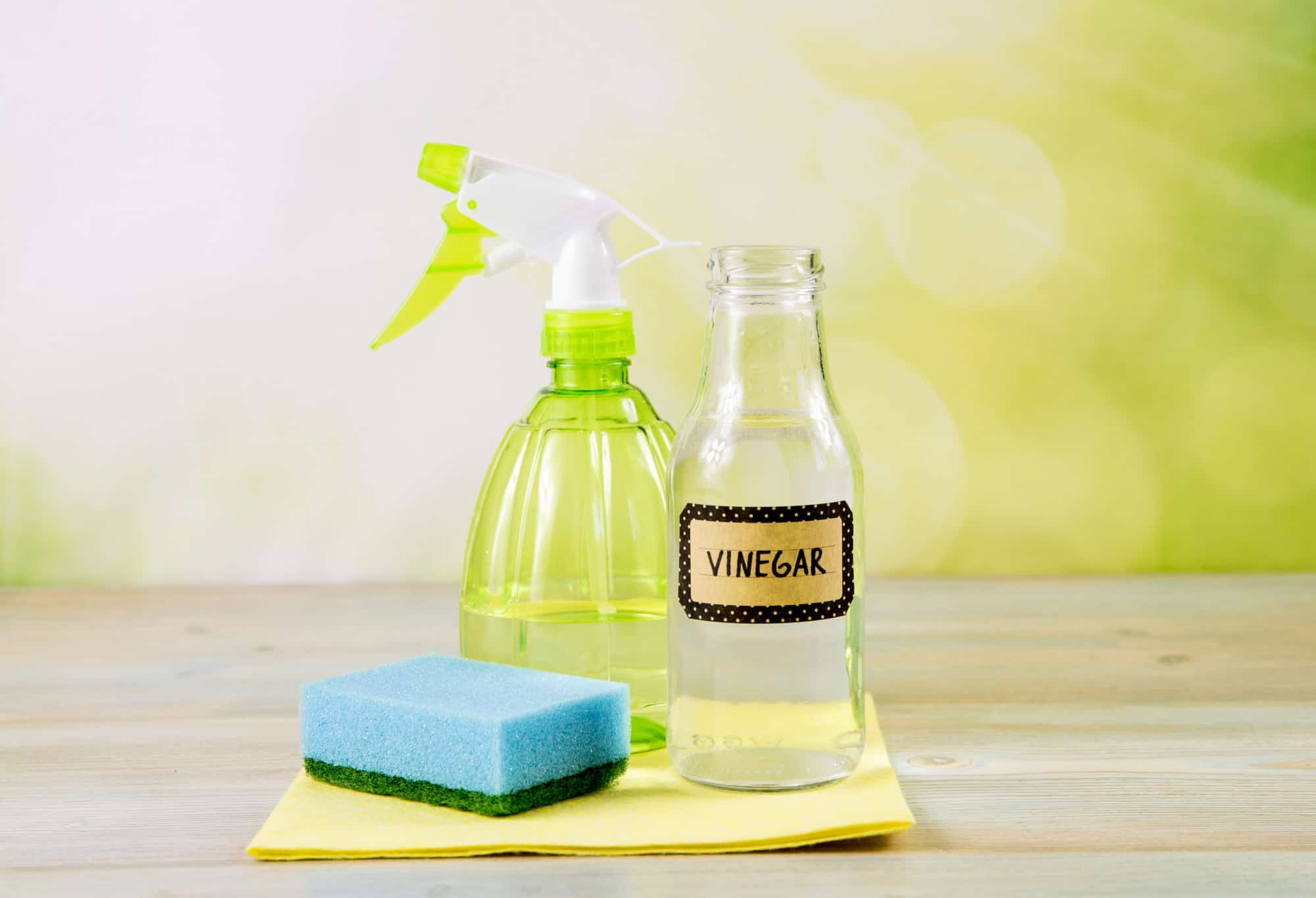 A spray bottle, a bottle labeled vinegar, and a sponge on a wooden table against a green background