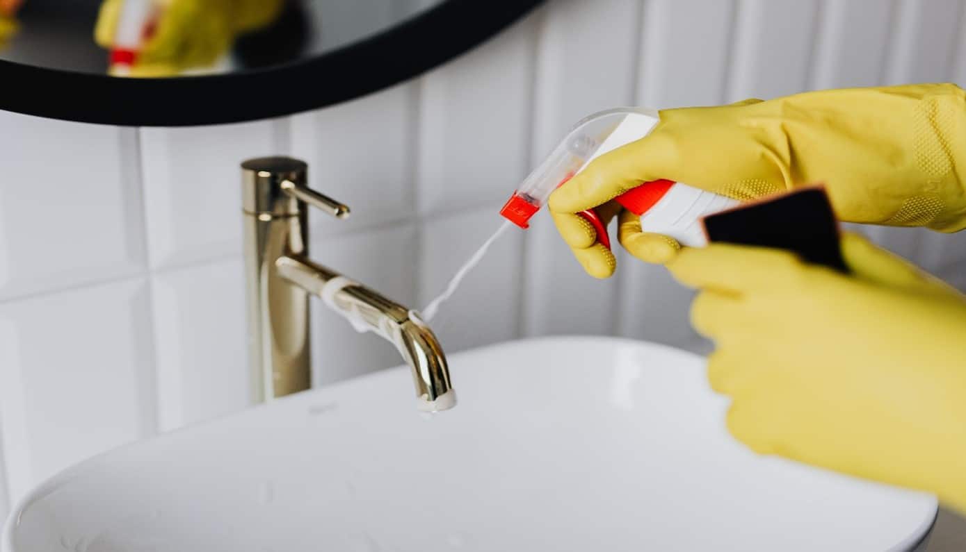 Person Cleaning Faucet with Disinfectant and Sponge.