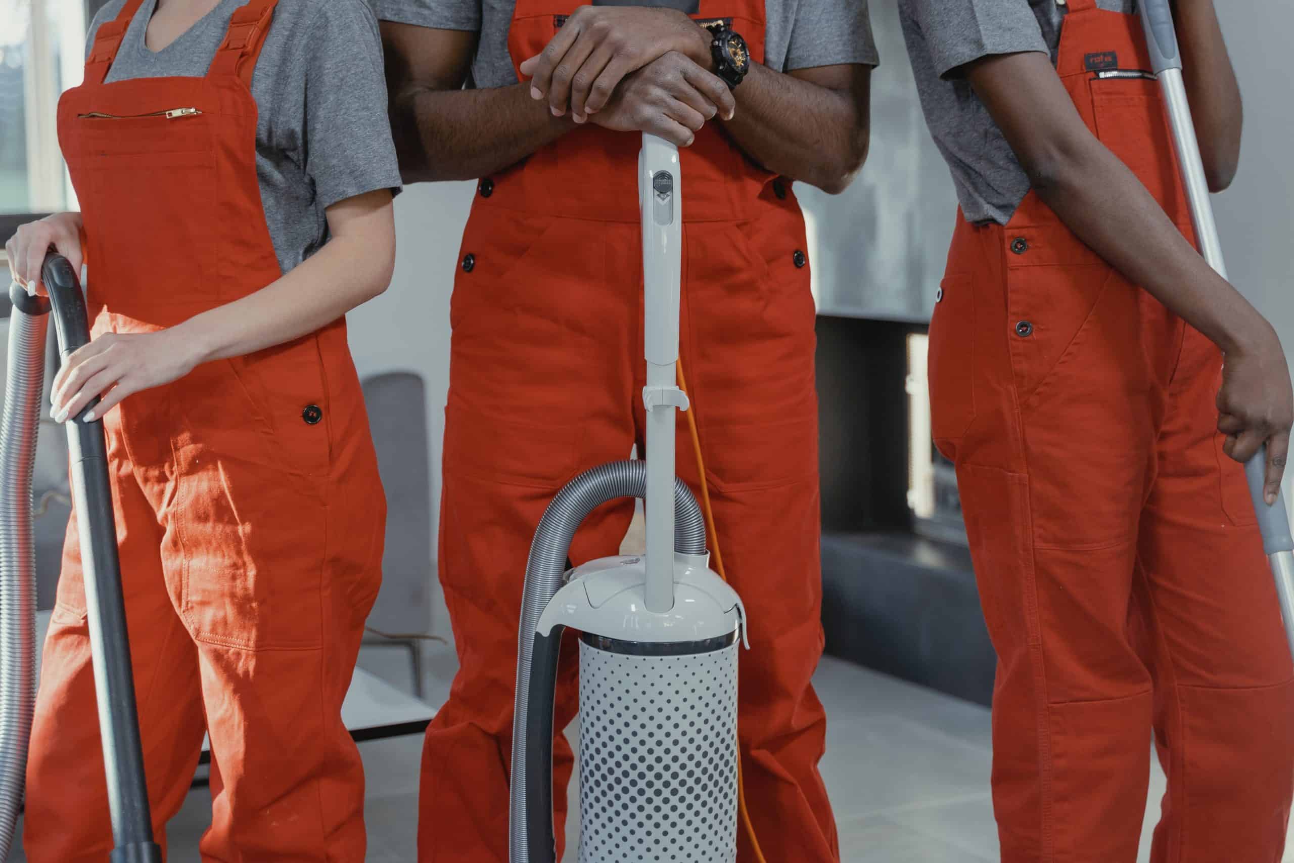 Three cleaners in orange overall uniforms each holding a vacuum.