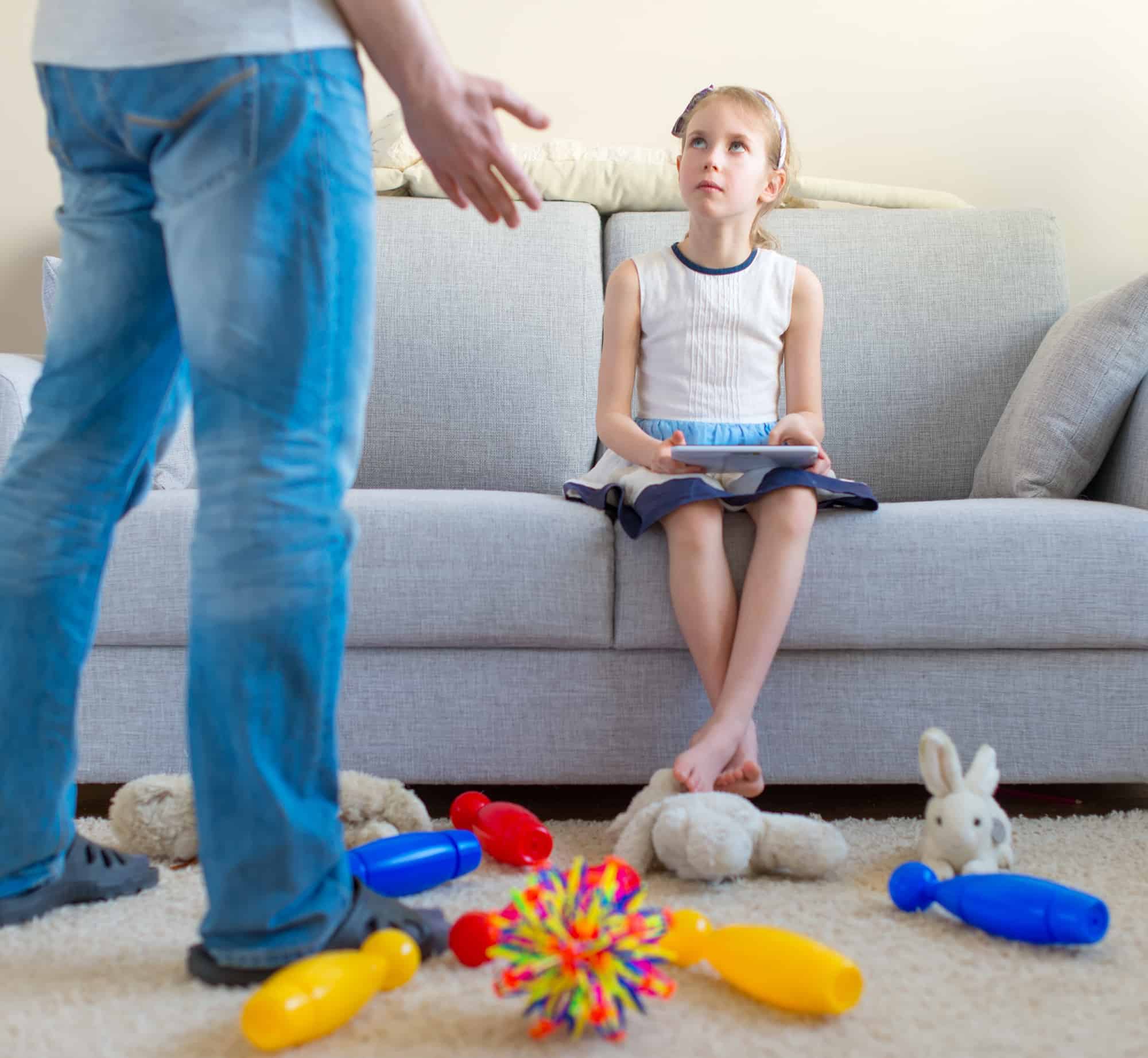 Girl sitting in couch with toys strewn about and ignoring her father.