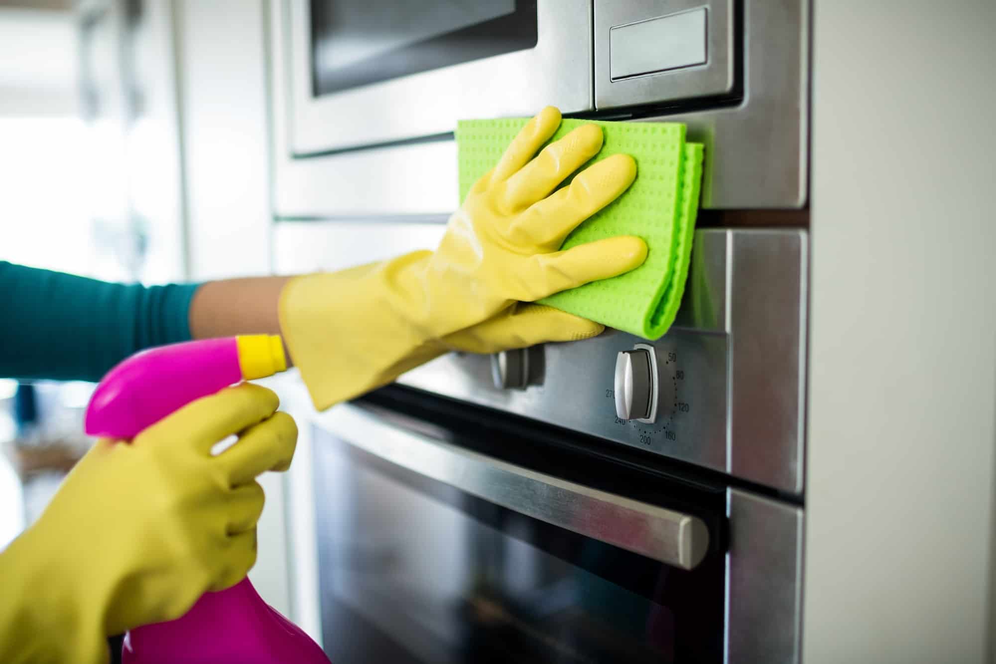 A pair of hands with yellow gloves on spraying and wiping down the stainless steel exterior of the oven and microwave.