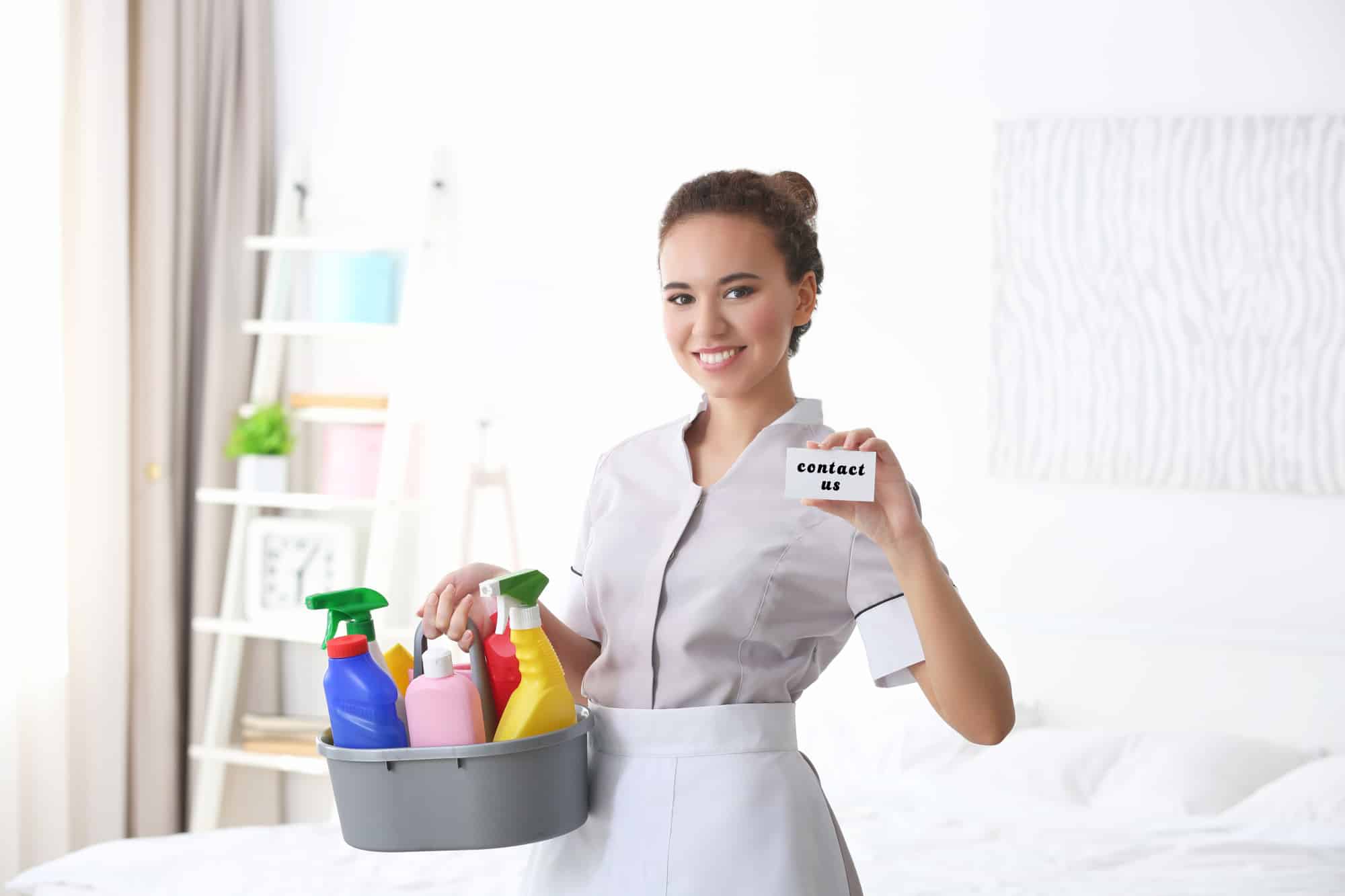Maid holding business card with text CONTACT US and cleaning supplies in bedroom.