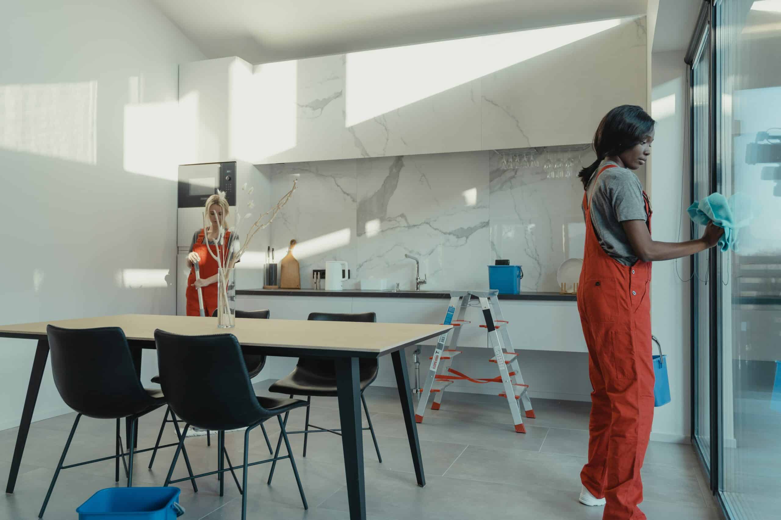 Two maids in orange outfits cleaning a kitchen.